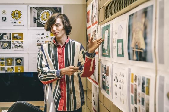 Photo of a male Chatham University student gesturing towards art hung on the wall and speaking